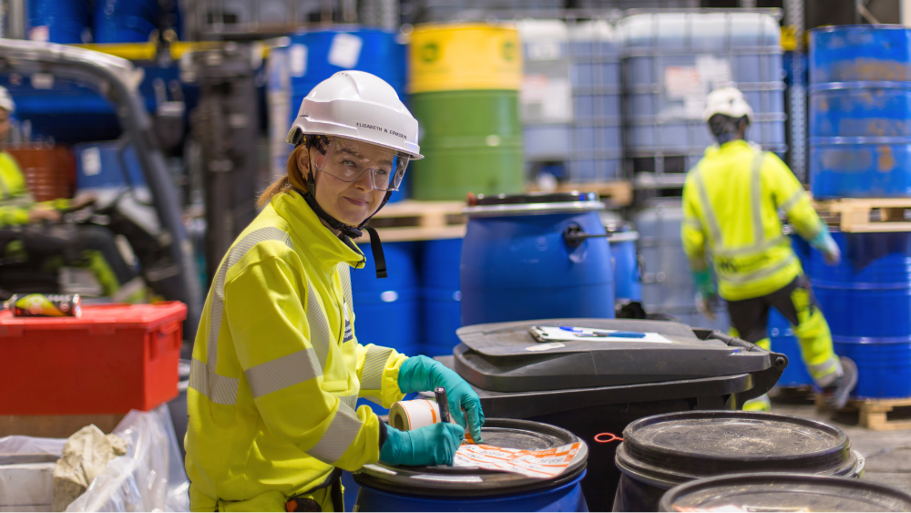 Kvinne i verneutstyr merker farlig avfall på beholder i industrihall.