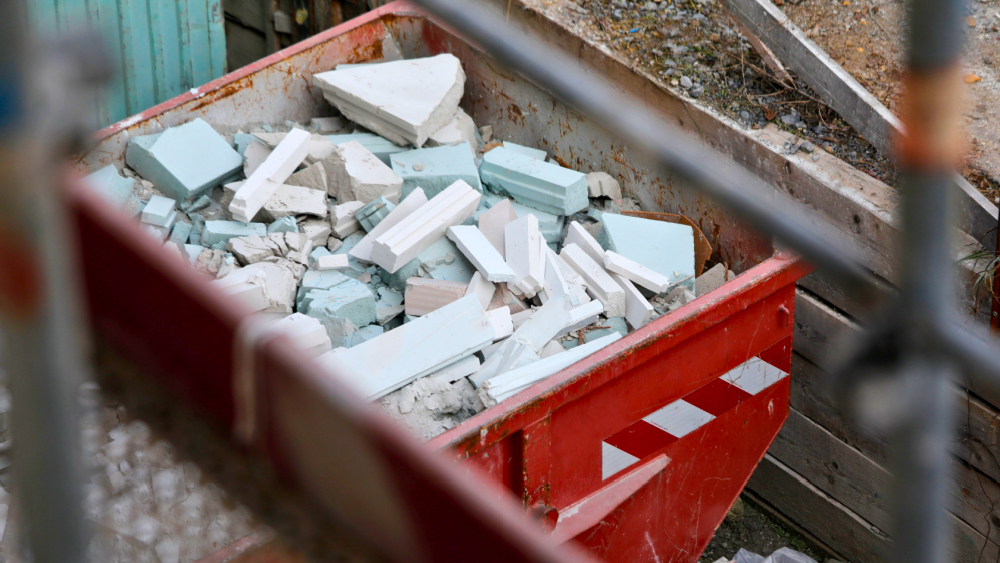 En rød container fylt med byggavfall, inkludert biter av gips og isolasjonsmateriale, på en byggeplass.