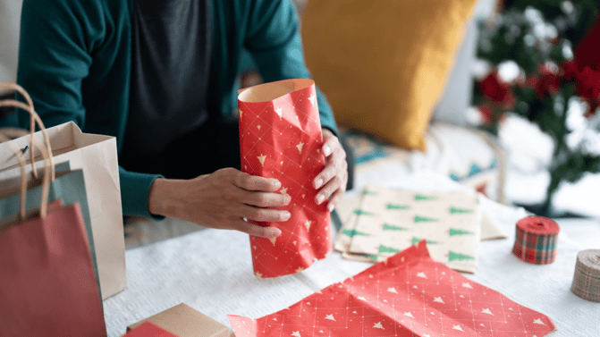Person pakker inn julegave med rødt papir ved siden av gaveposer, med et juletre i bakgrunnen.