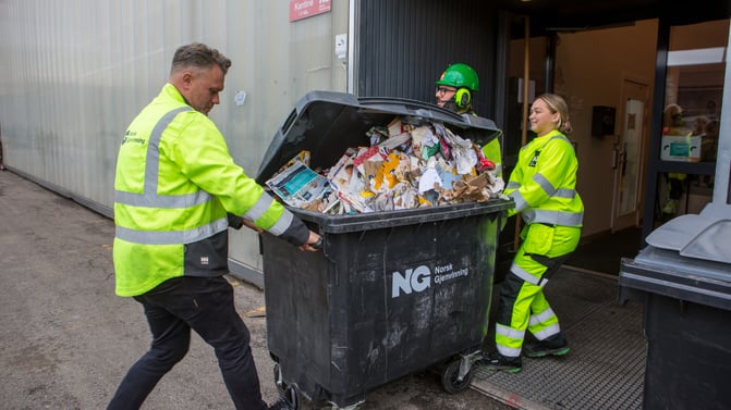 Tre NG-ansatte triller ut en full avfallsbeholder med papir og papp fra et bygg.