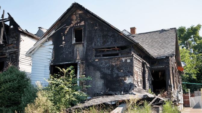 Utslitt, delvis brent enebolig omgitt av busker og trær.