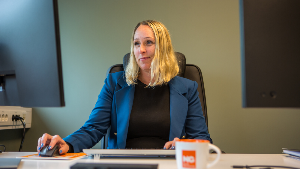 Kvinne ved kontorpult jobber foran to skjermer med NG-kopp og skjermer på pulten. 