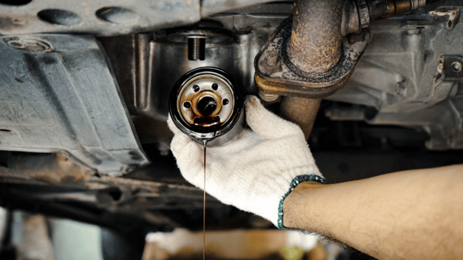 Oljefilter fjernes fra bilens underside av en person med hansker – olje drypper ned under demonteringen.